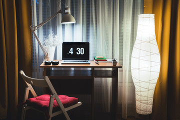 Modern evening interior of living room with small office desk and floor lamp against  window. Laptop on table ready to start late night work overtime. Working from home concept