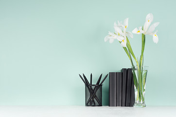 Spring soft students workplace with black stationery, books,  fresh white iris in trasparent glass vase on light white wood table and green mint menthe wall, copy space.