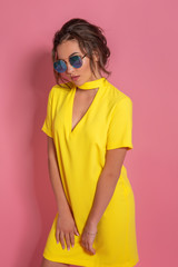 Beautiful girl wearing yellow dress and sunglasses posing on pink background in studio. Looking away of camera.