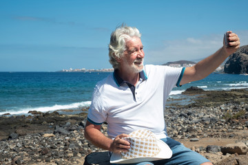 Smiling senior man using smartphone for a selfie on the pebble beach. Blue sky and sea behind him. Healthy lifestyle. Happy retirement concept