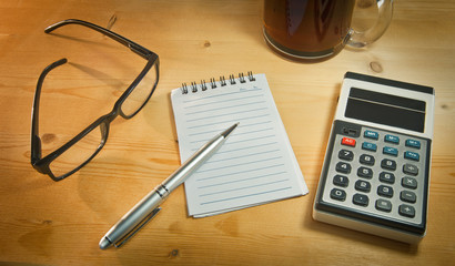 an old calculator black glasses a notepad a pen and a cup of coffee on a yellow wooden board