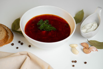 Beetroot soup in white bowl on a white table served with napkin, garlic, pepper, bread and saucy with sour cream. Traditional ukrainian, russian soup borscht with greens.