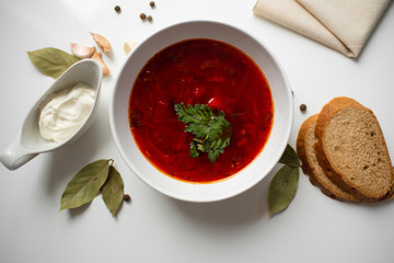 Beetroot soup in white bowl on a white table served with napkin, garlic, pepper, bread and saucy with sour cream. Traditional ukrainian, russian soup borscht with greens. Top view.