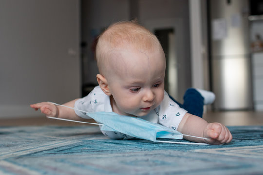 White caucasian baby boy playing with a face mask trying to take it of while staying at home on isolation and quarantine during pandemic of coronavirus virus covid-19 trying to wrip the mask