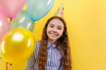 Fototapeta na wymiar Young girl in birthday party with colorful inflatable ballons isolated on a yellow background, little beautiful girl wearing birthday cap, smiling with happy face