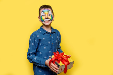 Portrait oh little happy boy in casual shirt with aquagrim on face, holding present box isolated on yellow background