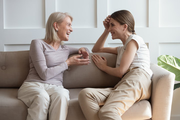Overjoyed middle-aged mother and adult daughter sit rest on sofa in living room have fun talking,...