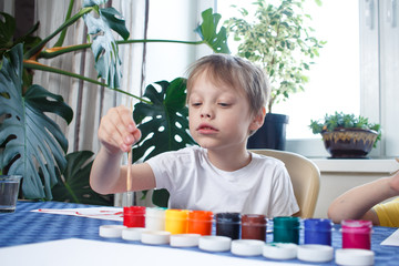 Happy cheerful child drawing with brush, in home interior during coronavirus self-isolation and quarantine. Creativity concept. A six year old boy with gouache.