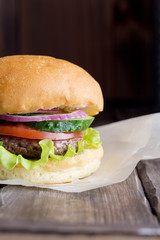 Burger on wood board at dark background. Fresh bun with beef, lettuce, tomato, cucumber and onion