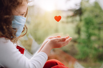 Cute small girl wear face blue mask sitting on sill near little heart as a way to show thank your...