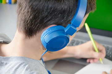 The boy is sitting in front of the computer with headphones on. The student holds a pen and writes...