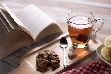 Cup of tea on wooden tray with cookie and lemon slices with open book in a cozy, relaxing and peaceful atmosphere