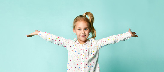Little blonde girl with ponytail, in shirt with hearts print. She smiling, spread her hands, posing...