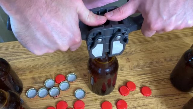 Craft beer brewing at home, man's hands closes brown glass beer bottles with plastic capper on wooden table with red crown caps. Close up horizontal HD video.