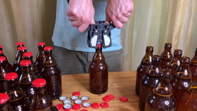 Craft beer brewing at home, man's hands closes brown glass beer bottles with plastic capper on wooden table with red crown caps. Close up horizontal HD video.