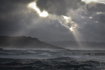 Big waves break on the coast, in the sunlit sea, passing through the clouds at sunset. Galicia, Spain.