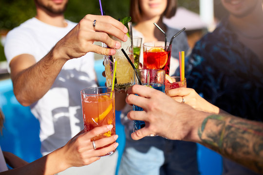 Close view of friends having fun at poolside summer party, clinking glasses with fresh summer cocktails near hotel swimming pool outdoors. People toast drinking juice at luxury villa on vacation.
