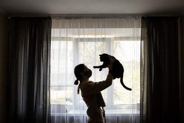 woman girl in a handmade protective mask, holds a black cat in her arms, stands at home near the window