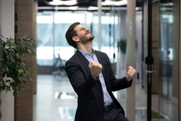 Millennial office worker in suit standing in hallway clenched fists feel overjoyed celebrate career...