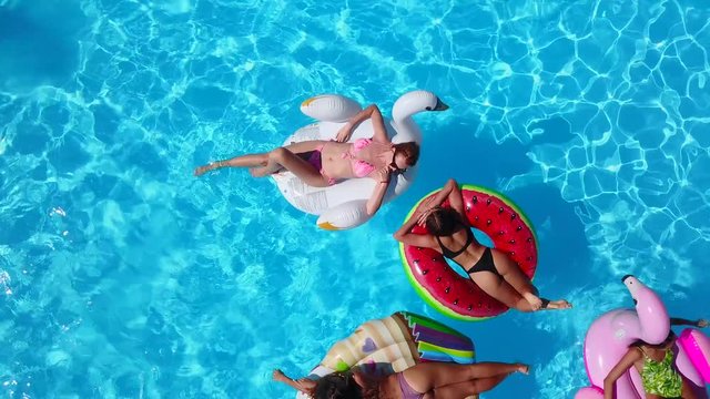 Aerial. Friends lay tanning on inflatable flamingo, swan, floaties and loungers. Happy young people bathe on air mattresses in luxury resort. View from above. Girls in bikini sunbathing in sun