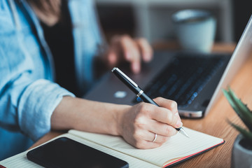 Young business woman sitting at desk, scrolling on laptop and taking notes in notebook. Close up of hand writing with pen - obrazy, fototapety, plakaty