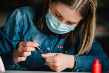 Woman doing manicure herself, draws, paints a brush, gel nail polish. Girl with protective mask makes a manicure at home. Home life during quarantine, coronavirus epidemic, pandemic, self-isolation.