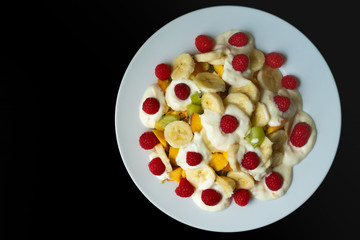 White plate with fruits, banana, raspberry, kiwi, apple and yogurt on a black background