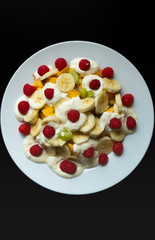 White plate with fruits, banana, raspberry, kiwi, apple and yogurt on a black background. Vertical photo