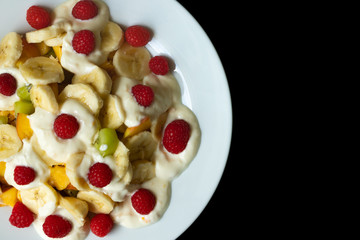 White plate with fruits, banana, raspberry, kiwi, apple and yogurt on a black background. Right space for copy