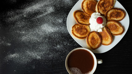 Pancakes on a white plate with raspberries and yogurt on a black background. Cup of coffee