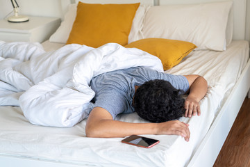 Young Asian man sleeping on the bed and turning off smartphone alarm clock in the morning. Tired...