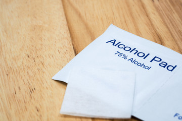 The close-up 1 unused alcohol pad over 2 alcohol pads in a package which are 75% alcohol with wood table background under warm light. © Sumate