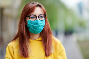 a girl with glasses, a yellow hoodie and a medical mask on her face walks down the street and looks away