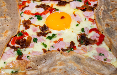 Crepe galette with ham, avocado, soft white cheese and poached egg on white plate.