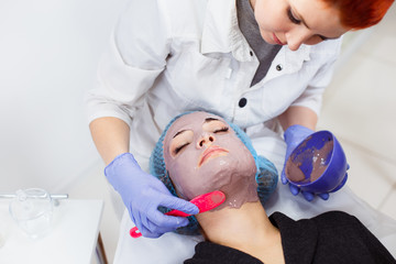 Naklejka premium The cosmetologist puts on the face of the client a mask in the beauty salon