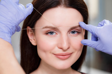 Cosmetologist plucks client eyebrows by thread. Close up