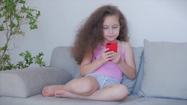 Curious cute girl preschool child using a digital tablet technology device looks telephone.Plays in the phone sitting on the sofa alone.Children tech addiction concept play game at home.