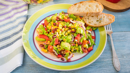 American-Mexican healthy salad with avocado, beans, corn, onions, lettuce and tomatoes in a plate with fresh rustic bread, top view