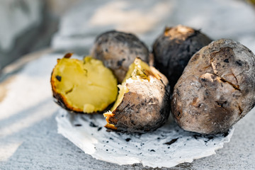 Baked in the fire potatoes in the skin according to the village recipe and method, directly in the ashes. Charred, fragrant tubers, a hot dish on a white stone plate