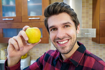 Young handsome happy man with a big smile showing an apple in the kitchen wearing a shirt 