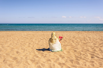 Back view of lonely Muslim woman  traveler in a colorful headscarf, scarf holding ripe of watermelon sitting on the sand on the beach and looking on the sea.