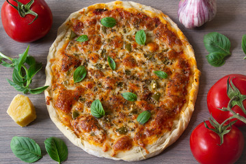 Homemade vegetarian margarita pizza with Parmesan and mozzarella cheese and traditional tomato sauce, topped with fresh green basil leaves on a wooden surface surrounded by its ingredients