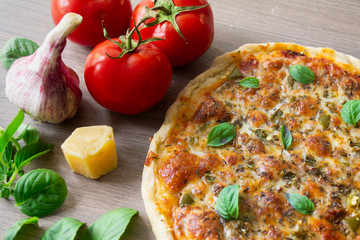 Homemade vegetarian margarita pizza with Parmesan and mozzarella cheese and traditional tomato sauce, topped with fresh green basil leaves on a wooden surface surrounded by its ingredients