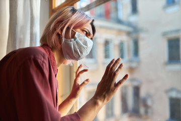 Young female patient with Covid-19 symptoms has to stay in hospital during quarantine, standing by window in disposable surgical mask, having stressed paranoid look, keeping hands on glass