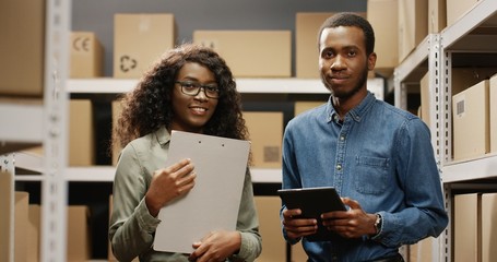 Male and female post workers looking straight to camera in postal storage of parcels and smiling in store.