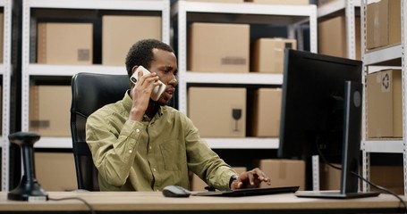 African American postman sitting at desk in postal store, working at computer and speaking on smartphone.