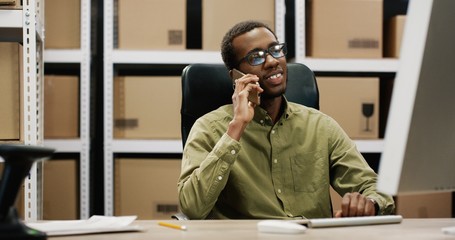 Fototapeta premium African American postman sitting at desk in postal store, working at computer and speaking on smartphone.