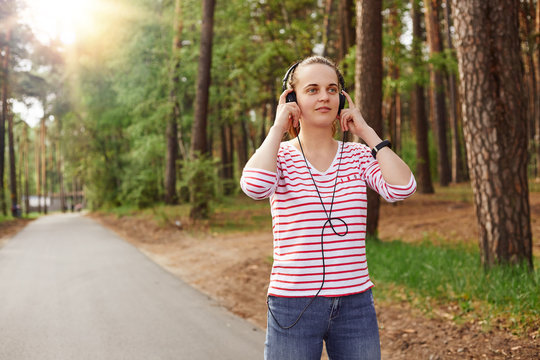 Horizontal photo of cheerful delighted good looking young female looking ahead, having headphones, listening to music, wearing jeans and sweatshirt, spending free time in peace. Rest concept.