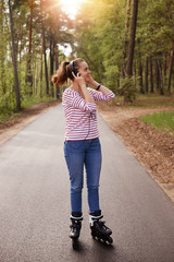 Outdoors shot of woman listening to music open air while spending time in forest, adorable female posing on asphalt road in wood, keeping hands on her headphones, looking aside, breathing fresh air.