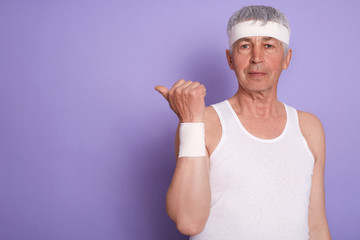Indoor shot of senior man wearing white t shirt and head band, mature male pointing aside with thumb while posing isolated over lilac studio background. Copy space for advertisement or promotion.